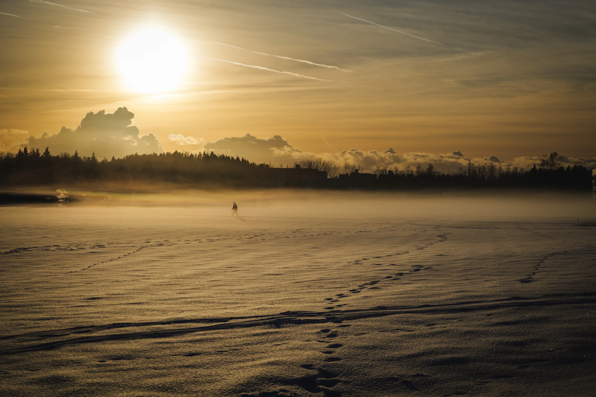 A lone person walks across a snow-covered field under bright winter sun, with low mist hanging over the landscape.
